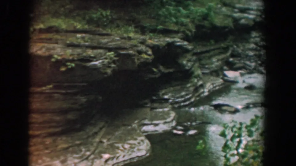 1958 Yorktown, Virginia 8mm frame of a shaded rocky stream bend with layered stone, shallow water, and foreground leaves.