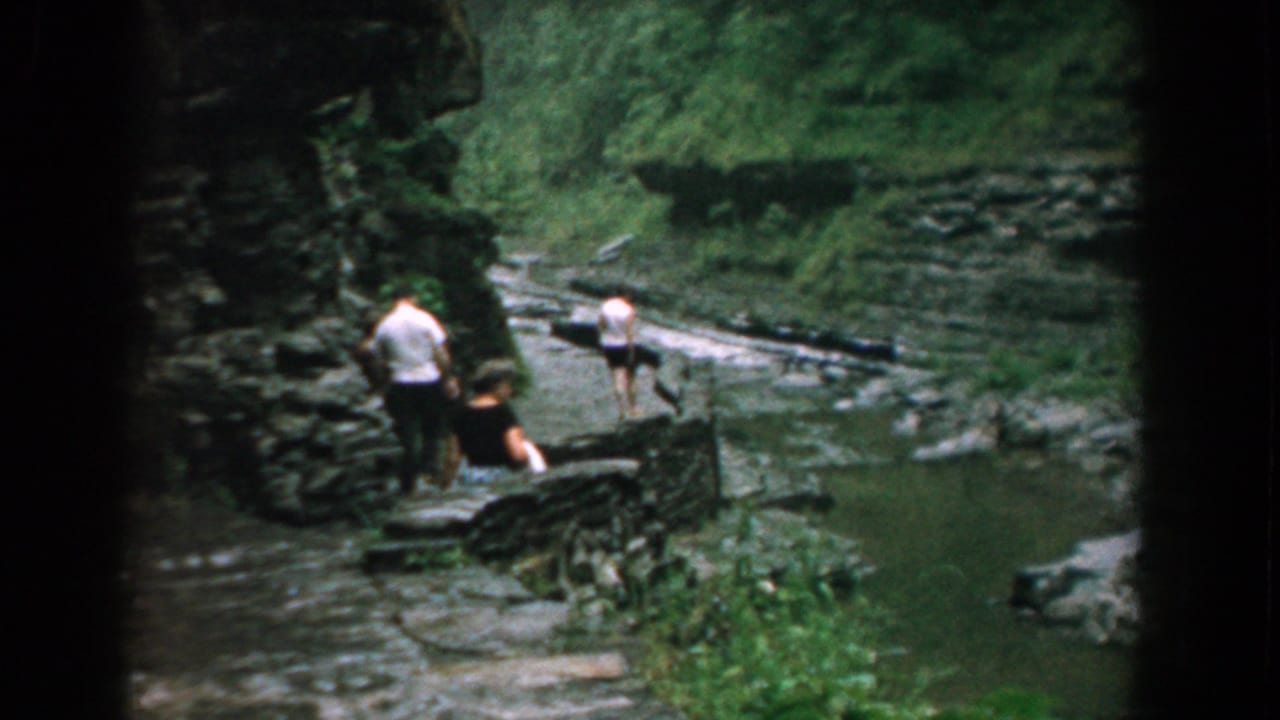 Three people walk along a dark stone path near a rocky stream in Yorktown, Virginia, in 1958 home-movie footage.