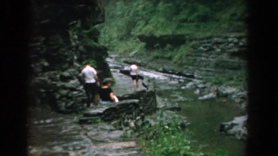 Three people walk along a dark stone path near a rocky stream in Yorktown, Virginia, in 1958 home-movie footage.