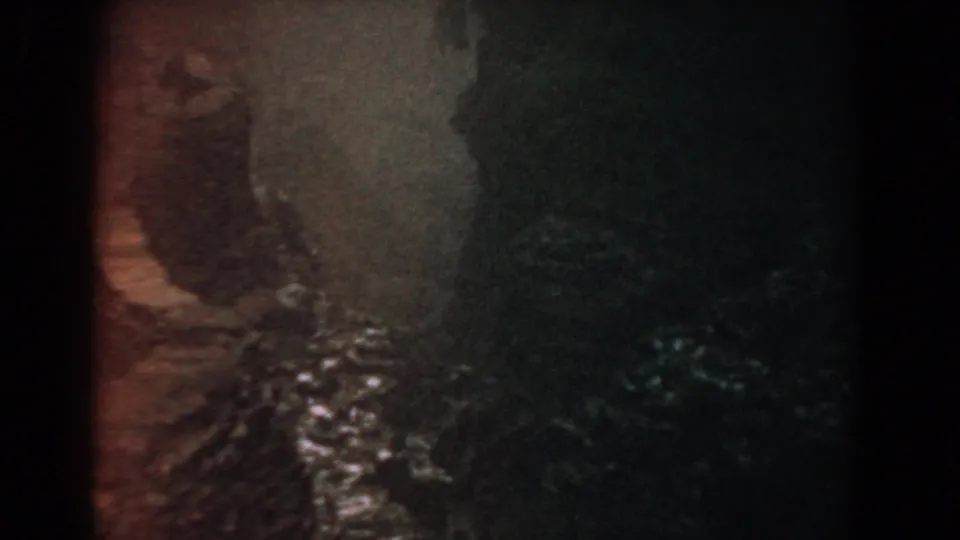 1958 home-movie frame appearing to show a narrow wooded path or riverbank with uneven terrain and bright water highlights.