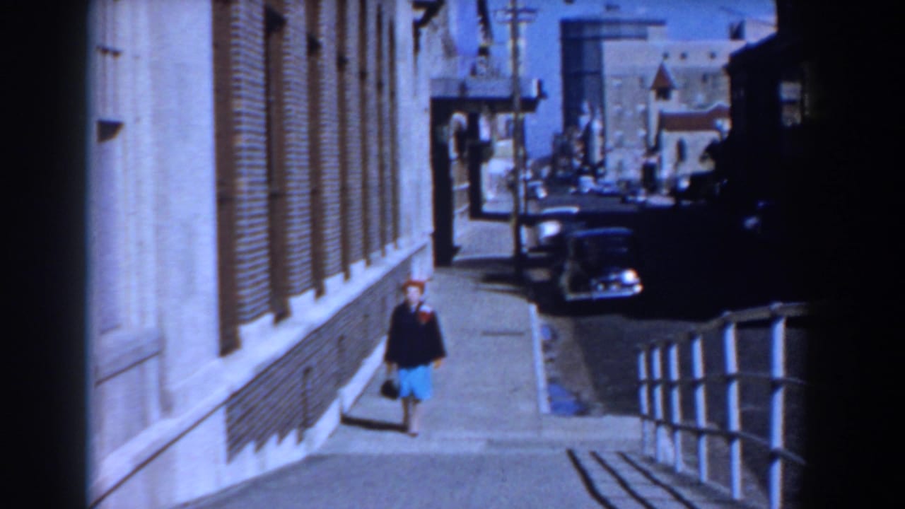 Woman in red hat and blue skirt walks along a sidewalk beside a brick building.