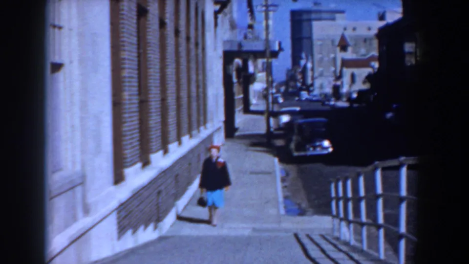 In Yorktown, Virginia, a woman in a red hat, dark jacket, and blue skirt walks alone beside a brick building in 1958 footage.