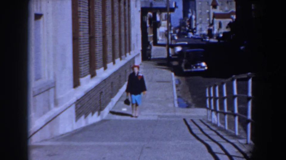 A lone woman approaches the camera on a Yorktown sidewalk, with mid-century parked cars and a brick facade in view.