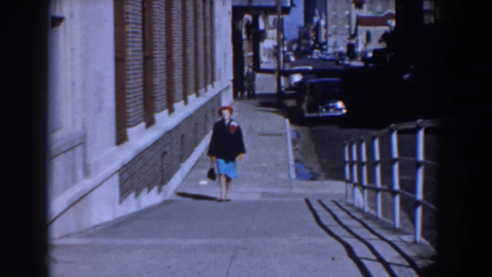 The 1958 Yorktown street view holds on the woman walking alone, with a brick building, parked cars, and railing shadows around her.