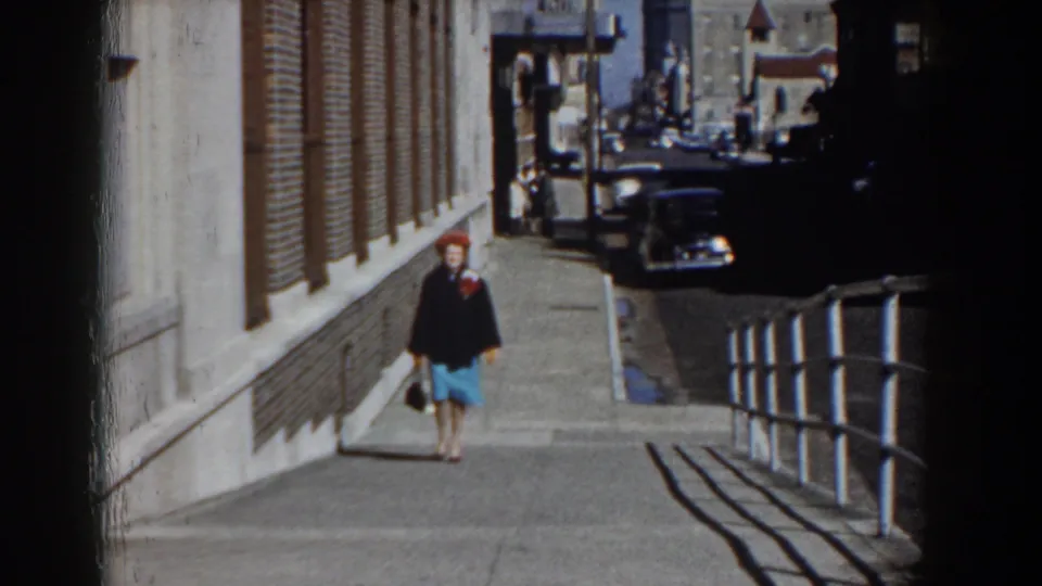 The woman continues uphill in Yorktown, Virginia, with mid-century cars and building facades visible behind her.