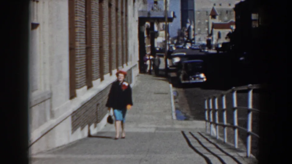 A closer 1958 frame shows the woman in a red hat, dark jacket, and blue skirt walking past railings and parked cars.