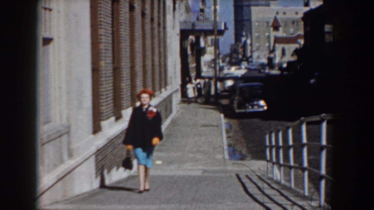 In Yorktown, Virginia in 1958, a woman in a red hat, dark coat, and blue skirt walks toward the camera on a sunny sidewalk.