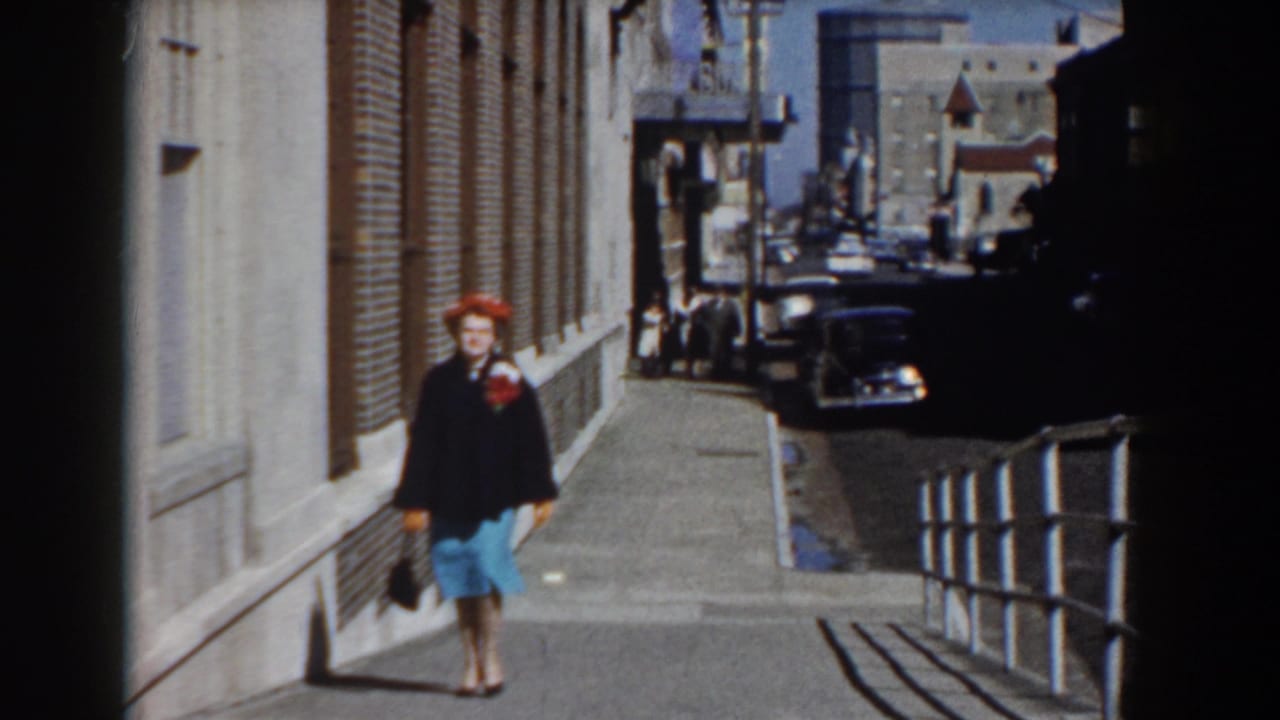 The woman continues toward the camera on a Yorktown sidewalk, framed by a brick wall, street railing, and parked 1950s-era cars.