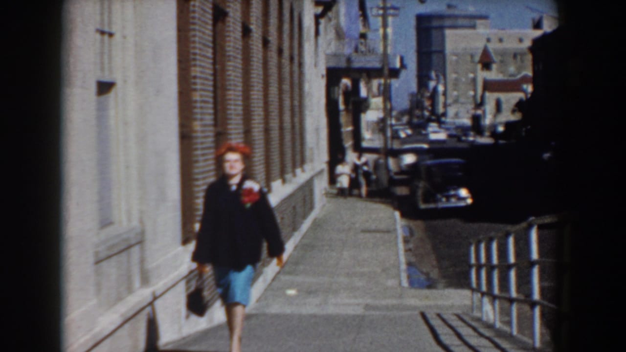 Closer view of the woman in 1958 Yorktown attire, carrying a handbag as she walks along the sidewalk past brickwork and parked cars.