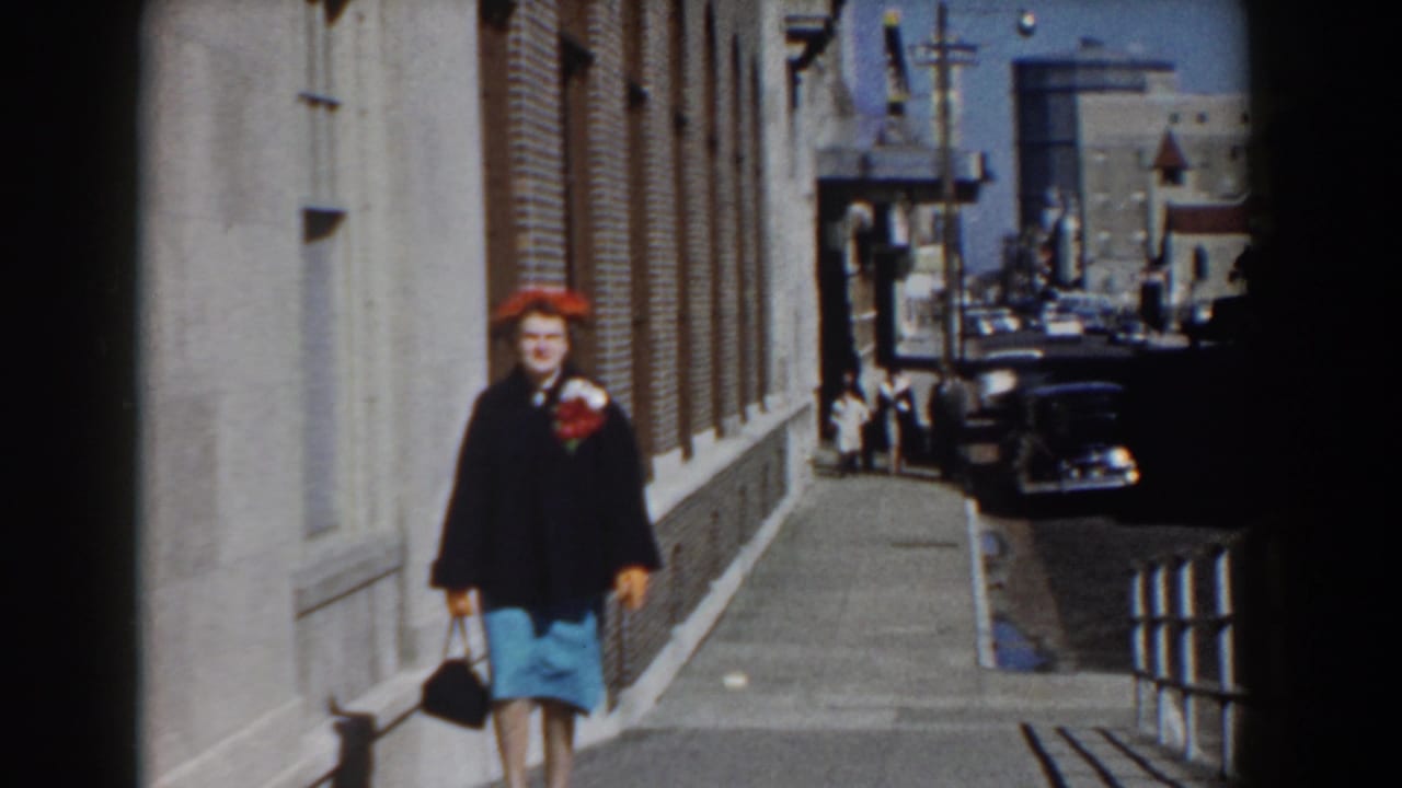 Near the end of the Yorktown street shot, the woman passes close to camera, with sidewalk railings and parked cars receding behind her.