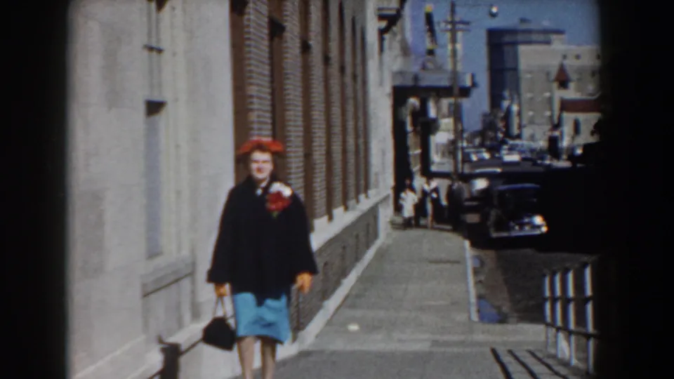 Near the end of the Yorktown street shot, the woman passes close to camera, with sidewalk railings and parked cars receding behind her.