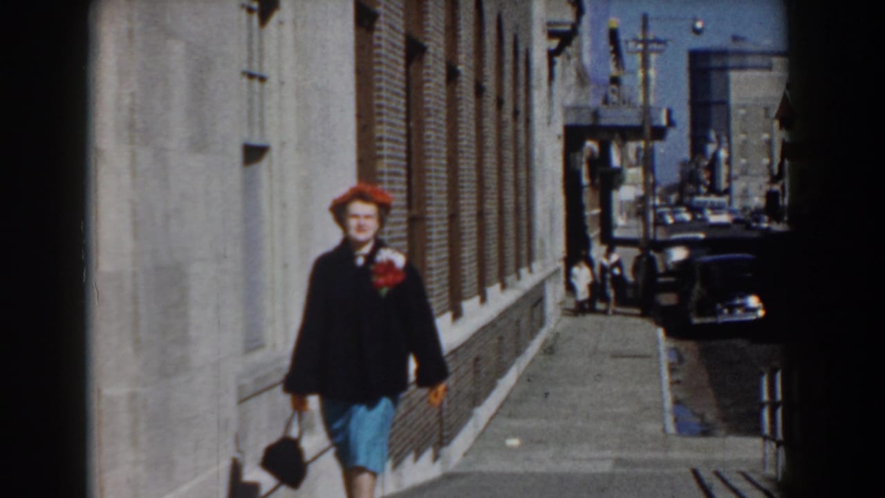 In Yorktown, Virginia, a woman in a red hat, dark coat, and blue skirt walks toward the camera on a bright 1958 sidewalk.