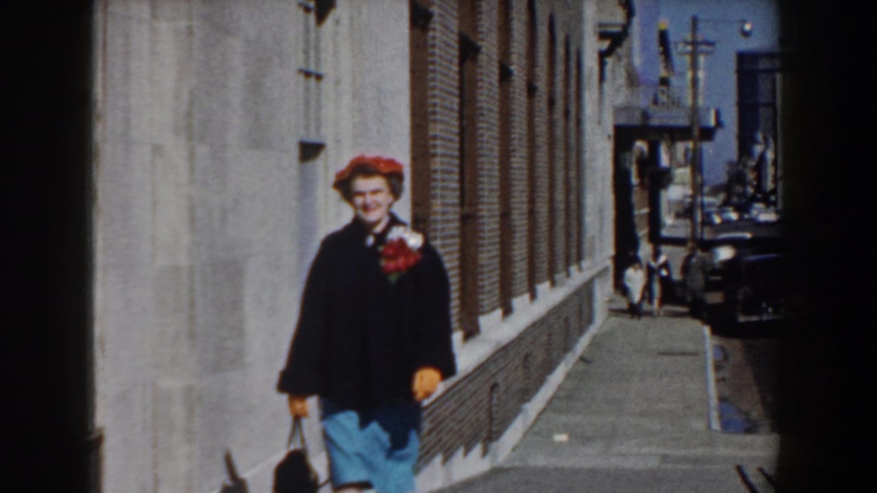 The woman smiles toward the camera as she passes brick buildings and parked cars on a Yorktown sidewalk in 1958.