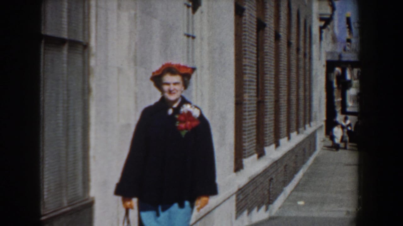 A closer 8mm frame shows the woman in a red hat, dark coat, and blue skirt walking beside brick buildings in Yorktown, Virginia.