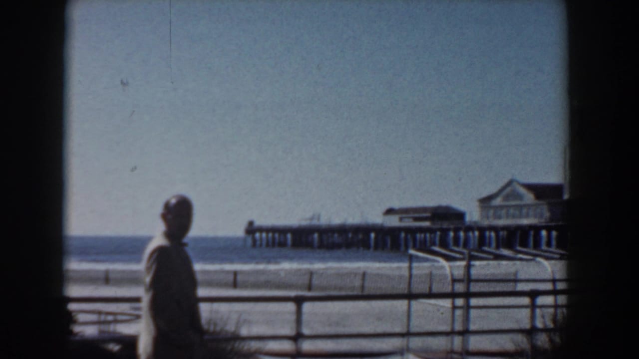 A silhouetted man stands near a waterfront railing, with sand, water, and a pier visible in the Yorktown-area 1958 home movie sequence.