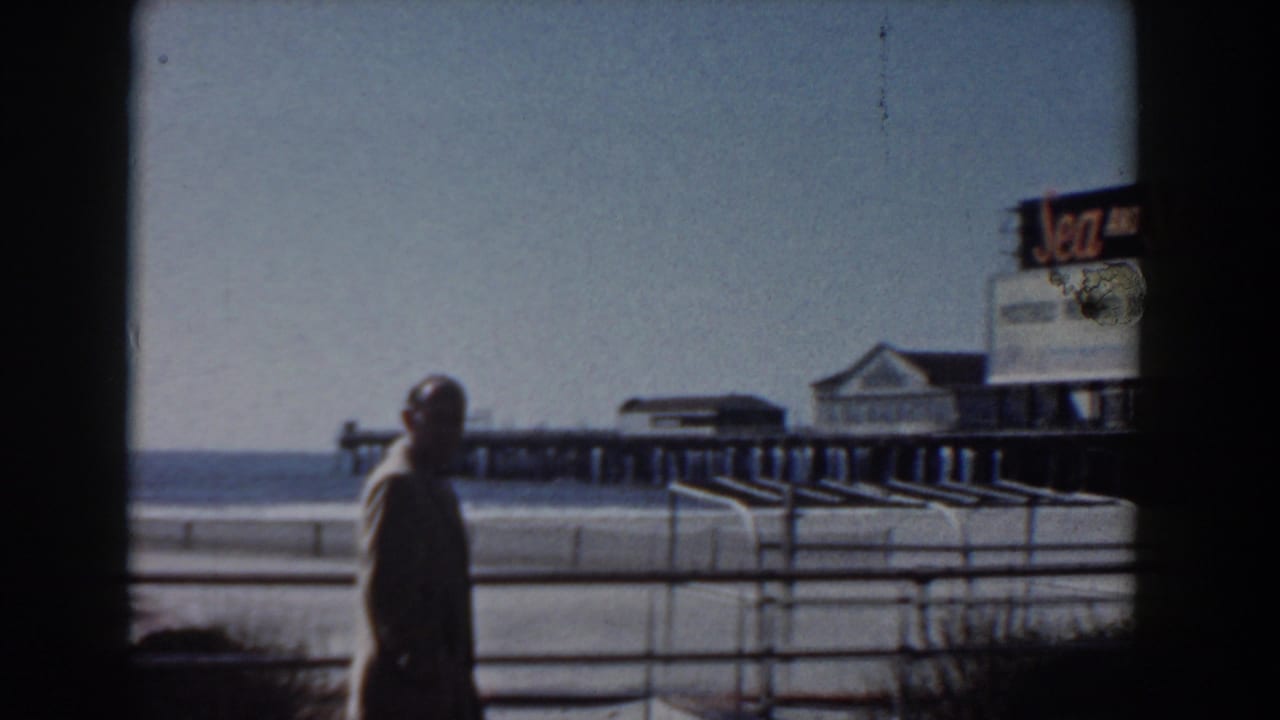 Man in a light suit walking beside railings with a pier and Sea and Ski sign in the background.