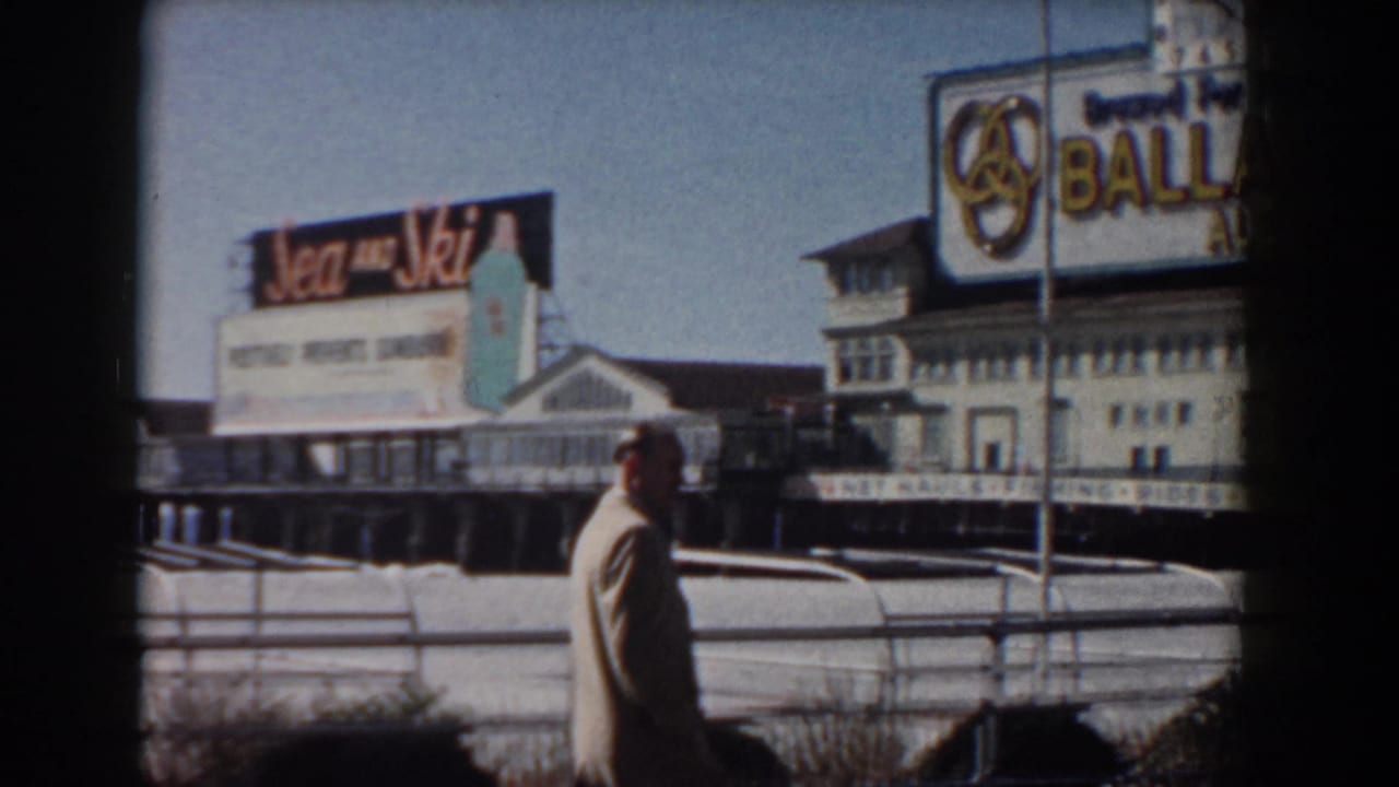 1958 waterfront boardwalk frame with a suited man walking past railings as Sea and Ski and Ballantine signage appear near the pier.