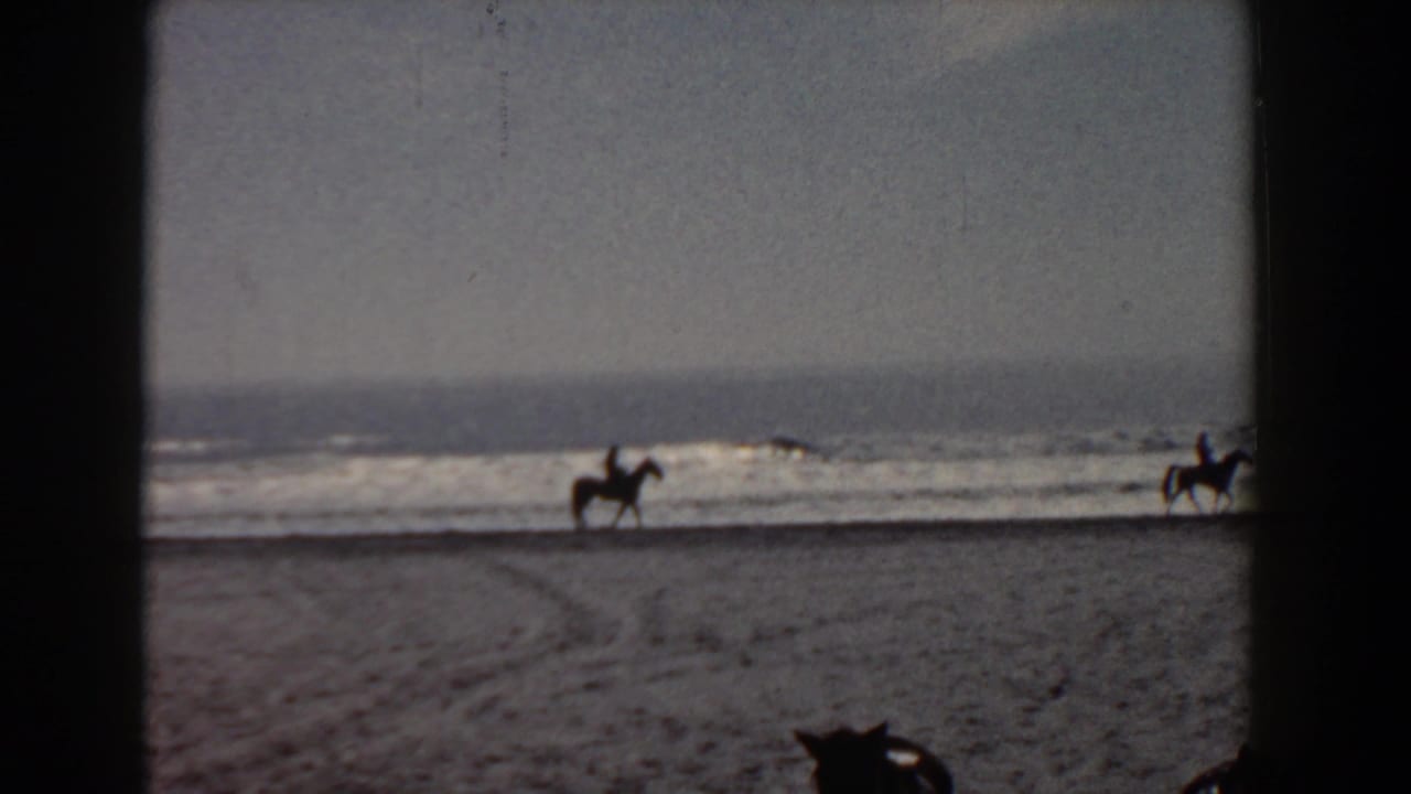 1958 Yorktown, Virginia home-movie frame showing two distant mounted riders on a sandy beach beside ocean surf.