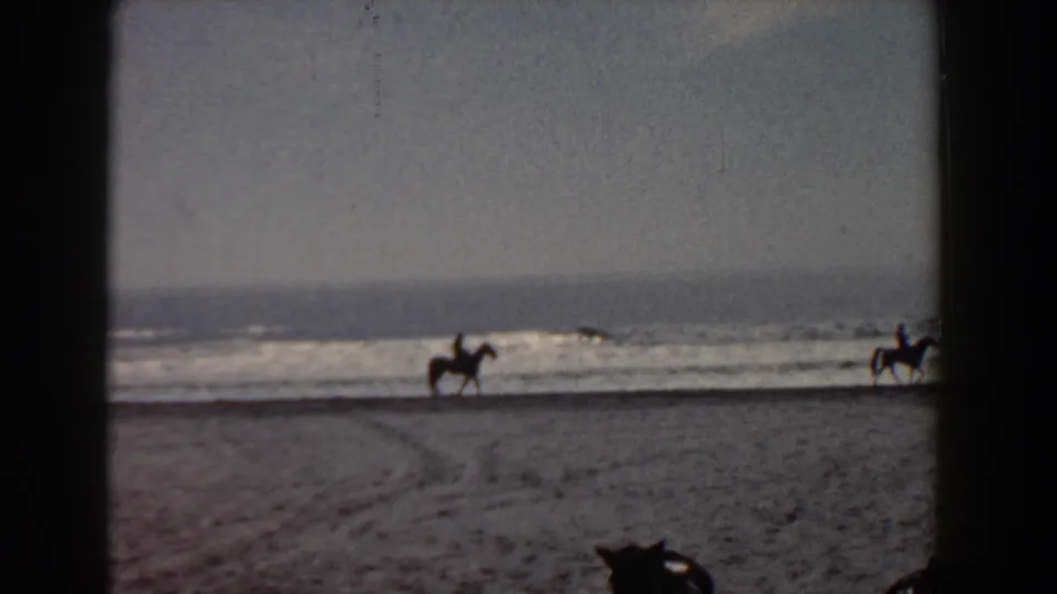 1958 Yorktown, Virginia home-movie frame showing two distant mounted riders on a sandy beach beside ocean surf.