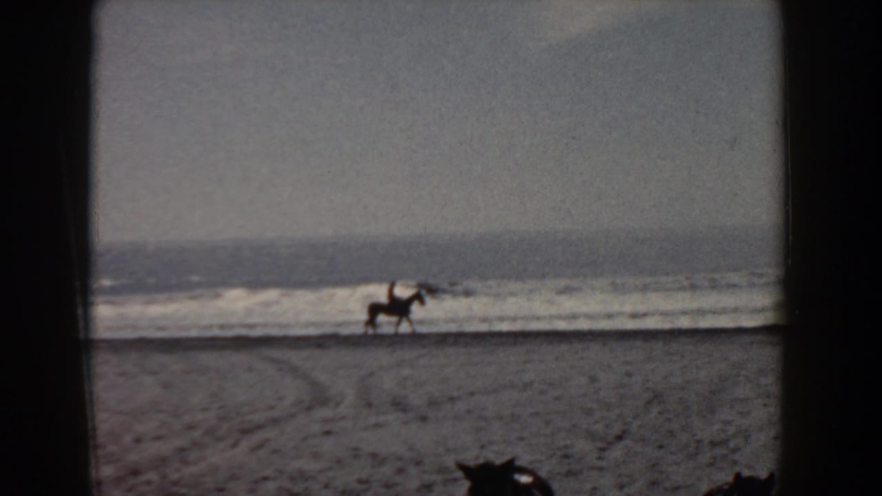 Distant horseback rider on a Yorktown beach in 1958, silhouetted against low waves and a hazy ocean horizon.
