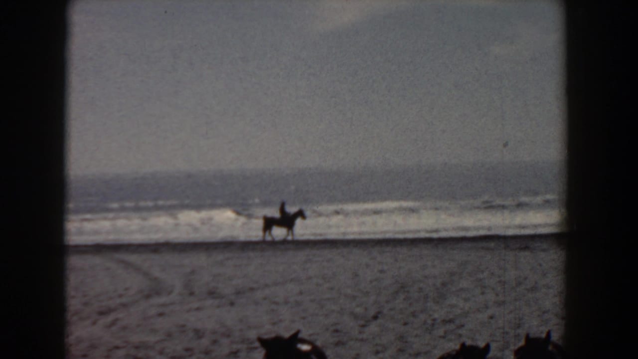Grainy 8mm frame of a distant mounted rider on the Yorktown beach, with waves and open sand filling the view.