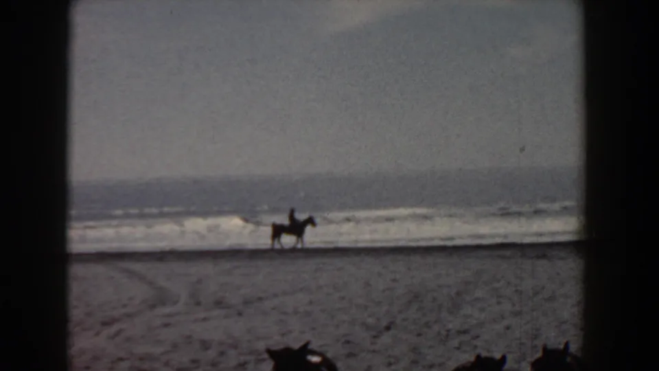 Grainy 8mm frame of a distant mounted rider on the Yorktown beach, with waves and open sand filling the view.