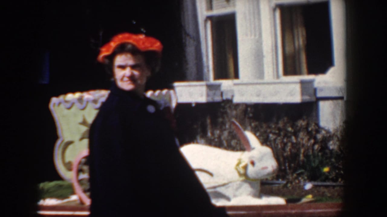 The woman remains beside the white rabbit statue, with house windows and garden planting behind her in the 1958 Yorktown footage.