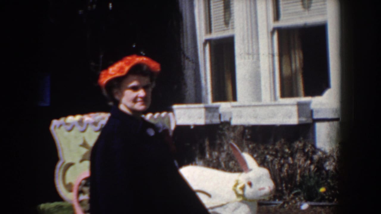 The woman turns toward the camera and appears to smile beside the large rabbit statue, with the house facade behind her.