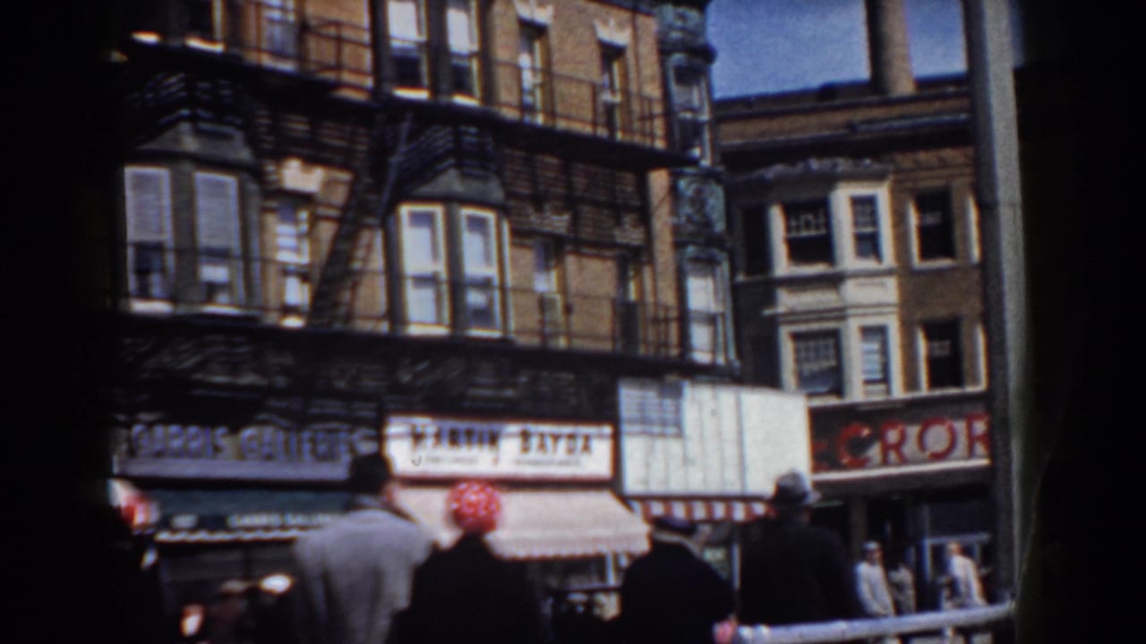 Pedestrians move along a commercial block with brick facades, awnings, and shop signs in supported 1958 Yorktown footage.