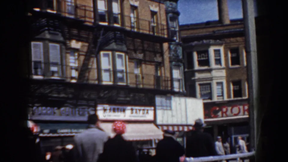 Pedestrians move along a commercial block with brick facades, awnings, and shop signs in supported 1958 Yorktown footage.