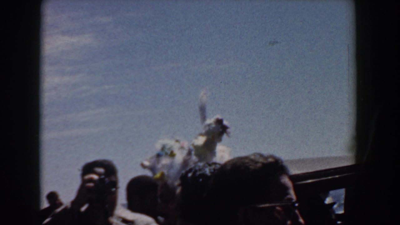1958 Yorktown home-movie frame showing spectators in hats and sunglasses near a feathered white costume by the water.