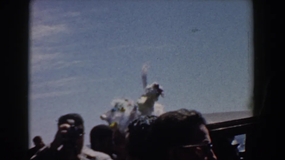 1958 Yorktown home-movie frame showing spectators in hats and sunglasses near a feathered white costume by the water.