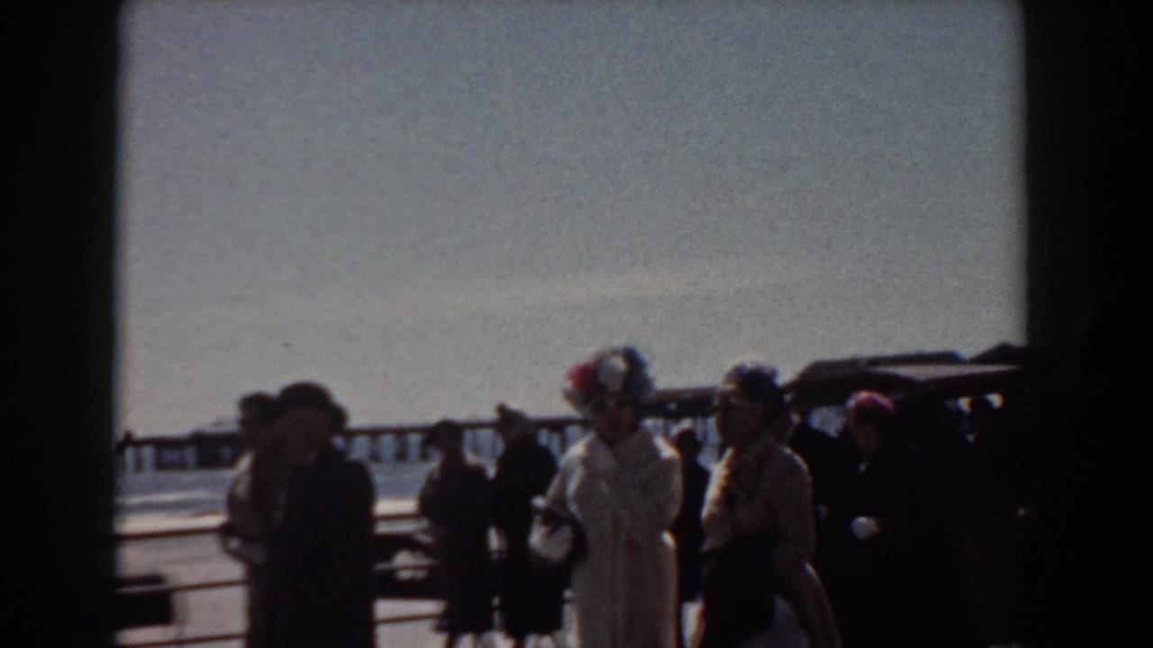 1958 Yorktown, Virginia home-movie frame showing formally dressed adults walking beside a waterfront railing with ocean and pier in the distance.