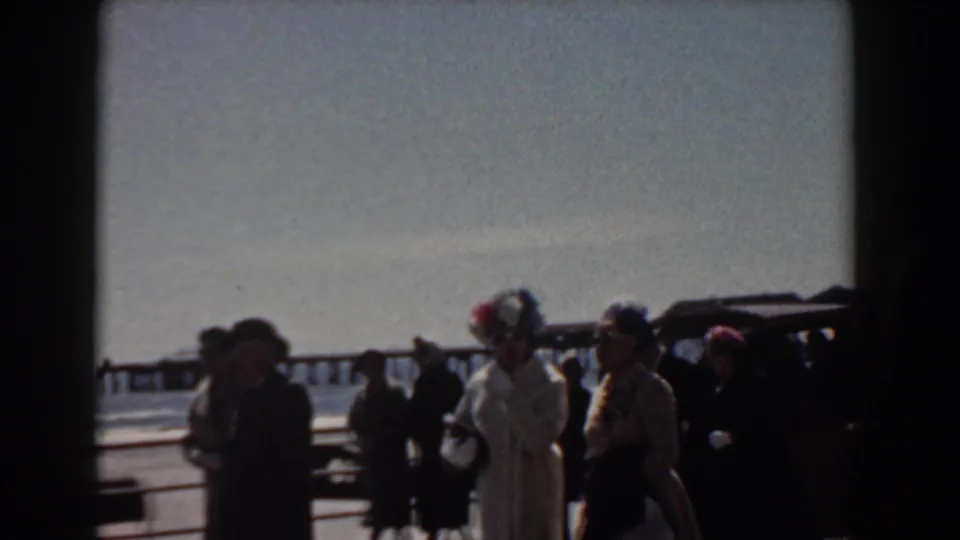 1958 Yorktown, Virginia home-movie frame showing formally dressed adults walking beside a waterfront railing with ocean and pier in the distance.