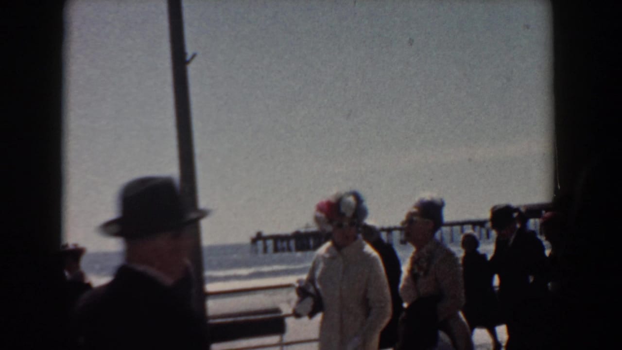 A 1958 Yorktown waterfront view shows adults in suits, coats, and decorated hats moving past the camera along the boardwalk.
