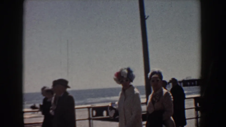 1958 Yorktown seaside footage shows adults in hats and coats moving through a tilted handheld frame near the ocean railing.