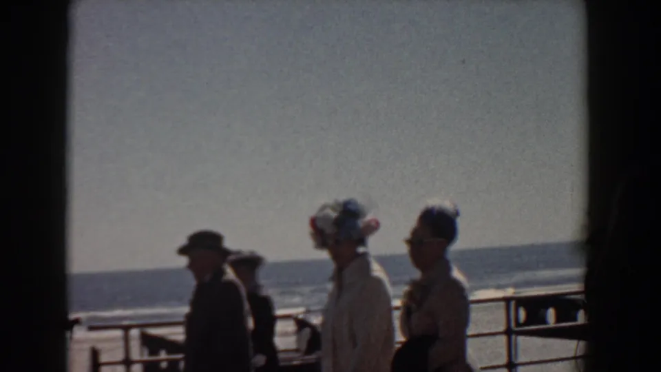 A Yorktown, Virginia 1958 home-movie still shows adults walking along a waterfront promenade under bright sky, with the ocean behind them.