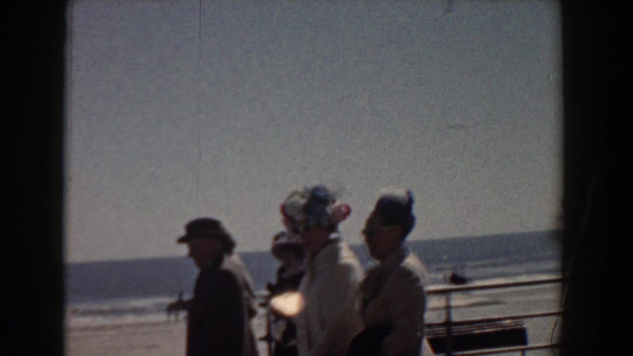 In 1958 Yorktown, Virginia, adults in mid-century hats and coats pass along a sunny waterfront promenade.