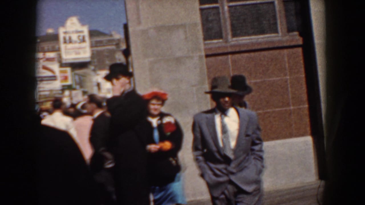 Adults in suits and hats pass a building facade near a public sign.