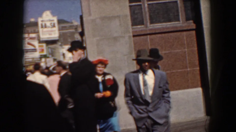 Suited men and women in hats move through a Yorktown waterfront walkway near a building and readable public sign.