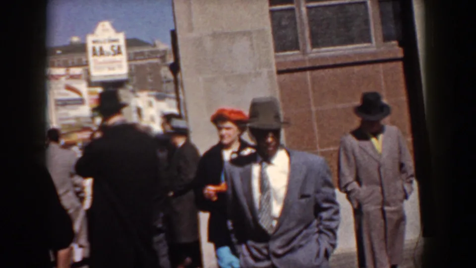 A suited man in a wide-brimmed hat walks toward the home-movie camera amid other Yorktown promenade visitors.