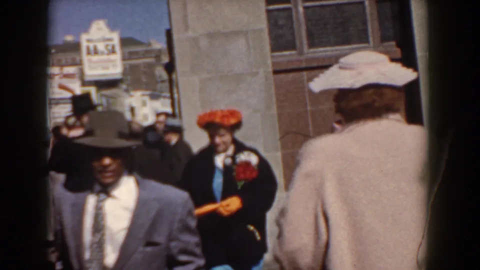 Near the end of the 1958 Yorktown clip, a suited man in a dark hat passes close to camera on a busy waterfront path.
