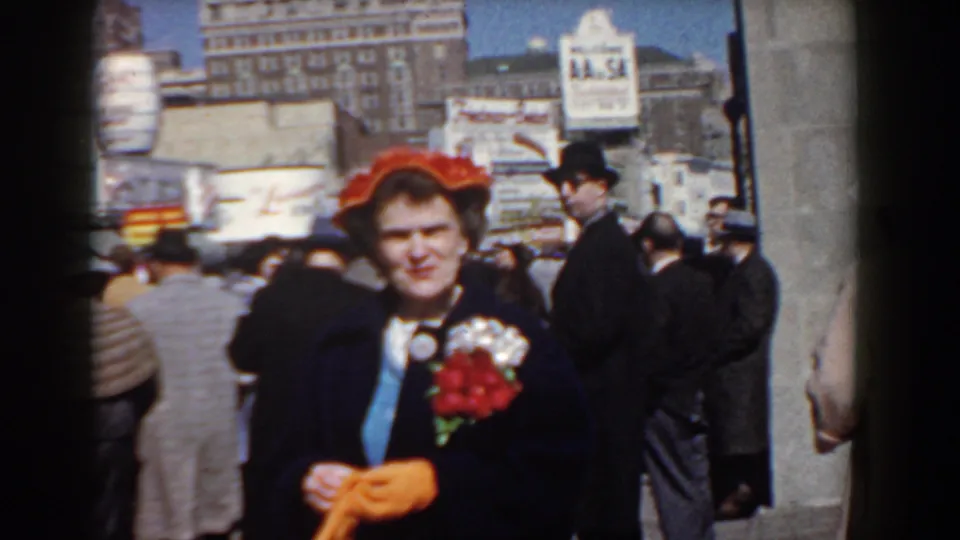 A woman in a vivid red hat faces the camera, holding red flowers and orange gloves amid a 1958 Yorktown sidewalk crowd.