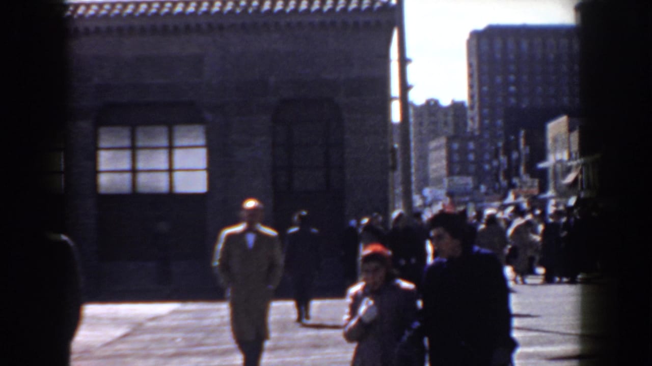 In this 1958 Yorktown street view, a man in a tan coat walks near a crowd with brick buildings and tall background structures visible.