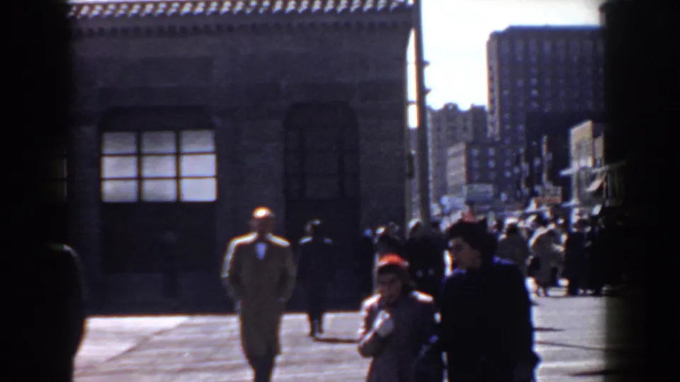 In this 1958 Yorktown street view, a man in a tan coat walks near a crowd with brick buildings and tall background structures visible.