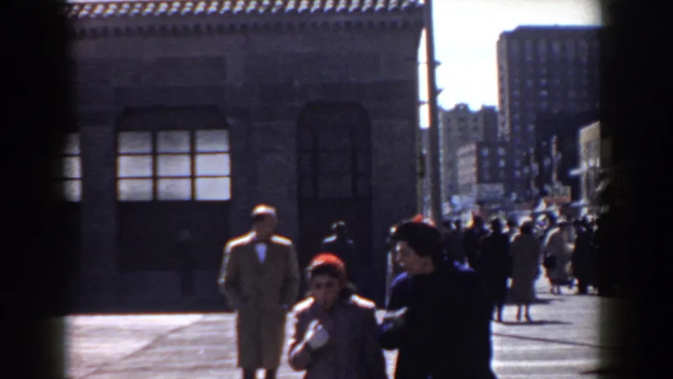 A 1958 Yorktown home-movie frame shows pedestrians in mid-century coats near a brick building and a crowded street beyond.