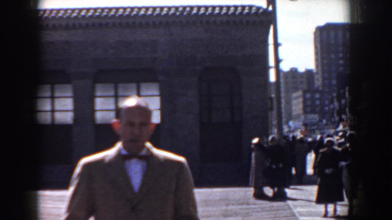 In Yorktown, Virginia in 1958, a man in a light-colored suit and bow tie approaches the camera beside a civic-looking stone building.