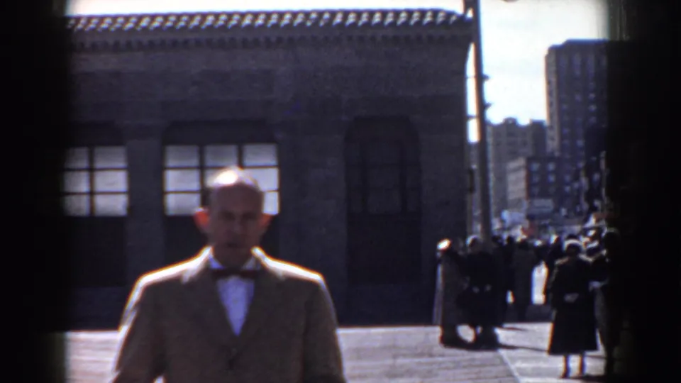 In Yorktown, Virginia in 1958, a man in a light-colored suit and bow tie approaches the camera beside a civic-looking stone building.