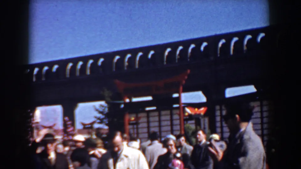 1958 Yorktown home-movie frame showing people near a dark-roofed storefront or pavilion with red posts and awnings.