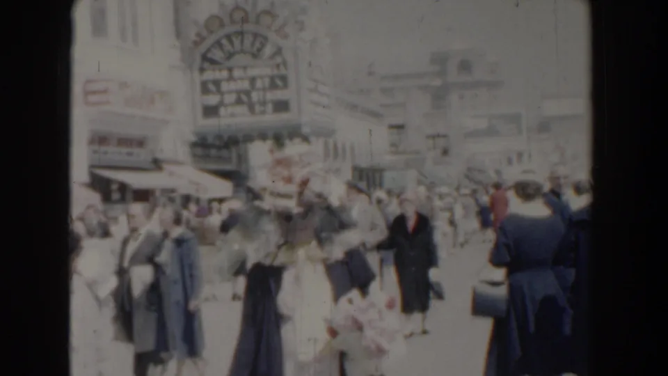 1958 Yorktown home-movie frame with men and women walking past shops, awnings, and a large theater-style sign.